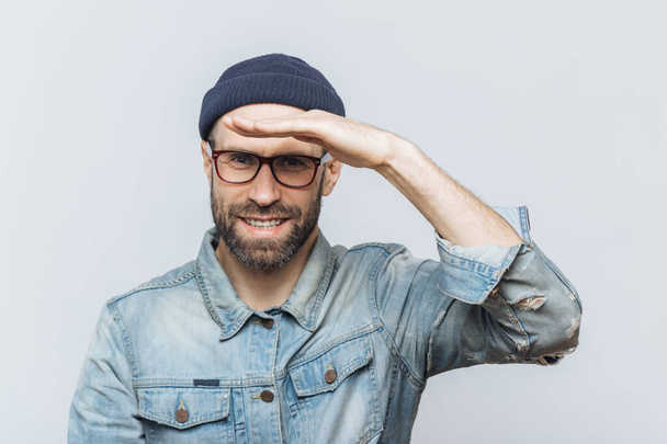 Cheerful unshaven middle aged male with thick stubble looks happily into distance, keeps hand near orehead, tries to see something, wears eyewear and denim jacket, isolated over grey background. - Photo, Image