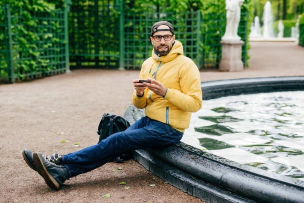 Sideways portrait of bearded male dressed in casual clothes wearing eyeglasses holding smartphone typing something looking aside having pensive expression isolated over fountain and monuments of park - Photo, Image