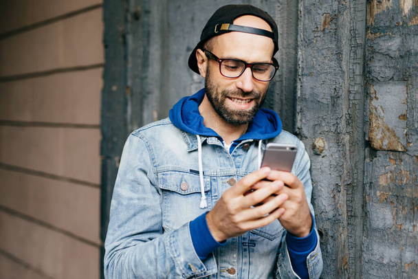 Ücretsiz Internet kullanımı, e-posta kontrol, komik video izlemek, uygulamaları smartphone tutan büyük gözlük takıyor denim anorak sakallı şık erkekte yakışıklı yatay portresi - Fotoğraf, Görsel