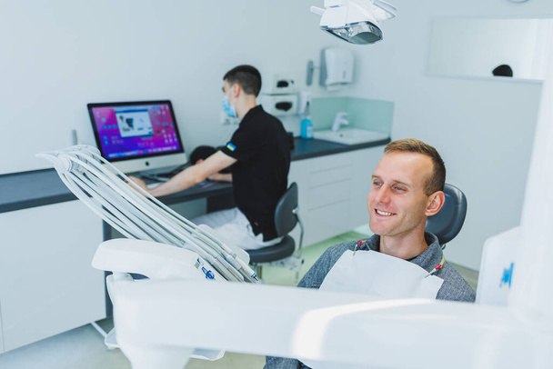 Mladý muž sedí v zubním křesle a zubař pracuje u počítače. Zubařská prohlídka - Fotografie, Obrázek