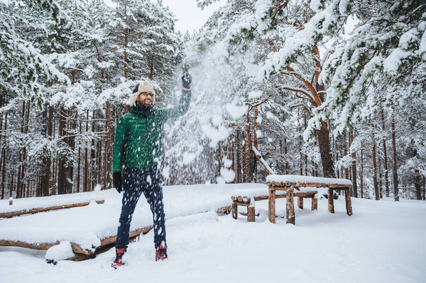 Outdoor shot of handsome bearded male dressed in warm clothes, has fun as throws snow in air, spends holidays in winter forest, expresses positiveness. Recreation and weather concept - Photo, Image