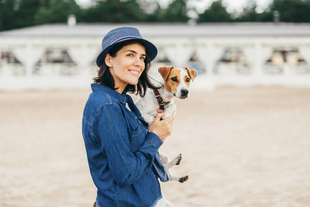 schöne Frau trägt modischen Hut und Jeanshemd, geht mit ihrem Hund im Freien spazieren, erkundet unbekannte Orte, blickt nachdenklich in die Ferne, hat glücklichen Gesichtsausdruck und ein angenehmes Lächeln - Foto, Bild