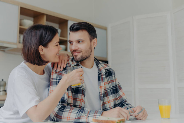 Lovely couple look at each other with love, woman holds glass of juice, pose together near kitchen table, have pleasant talk during breakfast time, enjoy domestic atmosphere. Happy family concept - 写真・画像