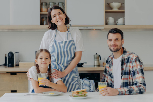 Positive friendly father, mother and their small daughter being in good mood, spend time together, pose at kitchen, gather to have breakfast, enjoy domestic atmosphere and day off. Time to eat - Photo, Image