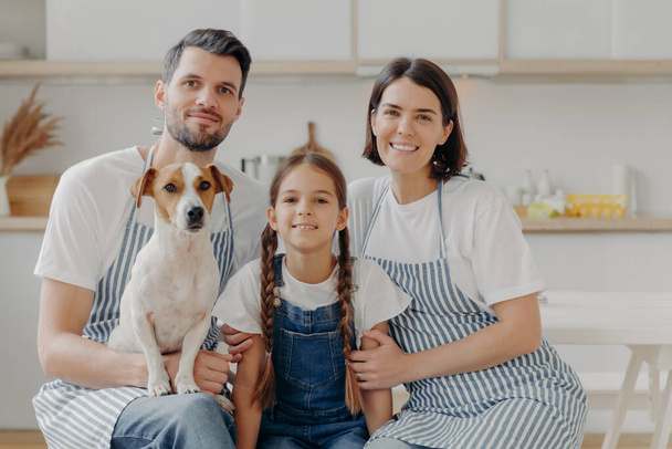 Family portrait of father, mother, daughter and pedigree dog pose together for making memorable photo, have rest after preparing dinner, poses against kitchen interior, have happy expressions - Zdjęcie, obraz