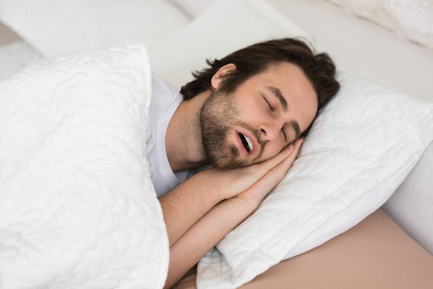Tired young caucasian male with open mouth sleeping on white soft bed in bedroom in spare time, enjoy quiet alone, copy space, close up. Snoring, rest and relaxation at weekend, healthy sleep at night - Photo, Image