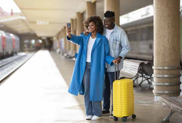 Sorrindo casal preto na estação ferroviária tomando selfie com Smartphone, felizes jovens afro-americanos cônjuges posando para câmera enquanto em pé na plataforma com bagagem, esperando por trem juntos - Foto, Imagem
