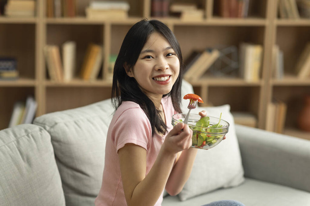 Концепція здорової веганської дієти. Щаслива азіатська леді сидить на дивані вдома з мискою свіжого овочевого салату, їсть смачний обід. Молода жінка насолоджується веганською їжею, пропагуючи чисте життя
 - Фото, зображення