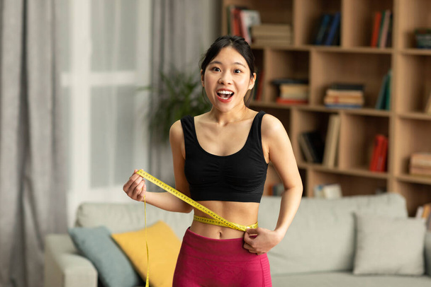 Porträt einer positiven Asiatin in Sportkleidung, die ihre Taille mit Klebeband misst und froh ist, zu Hause abzunehmen. Gesunder Lebensstil und Schlankheitskonzept - Foto, Bild