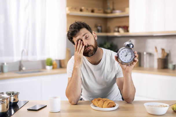 Unhappy tired sleepy millennial caucasian male with beard with alarm clock rubs his eyes at table, has breakfast alone in kitchen interior. Sleep deprivation, waking up early, overwork, time to work - Photo, Image