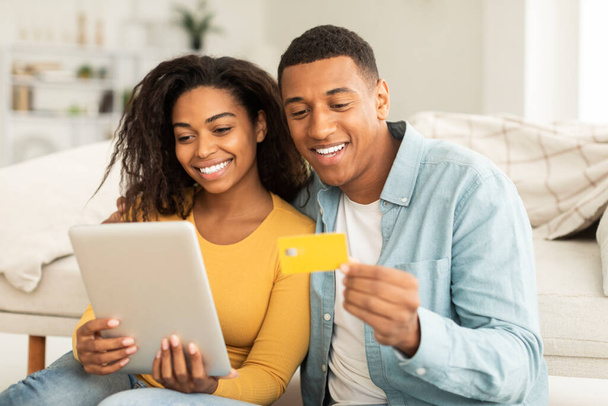 App for online shopping. Glad millennial african american guy hug lady, use tablet, credit card for order purchase, check banking in living room interior. Shopaholics enjoy sale and cashback at home - Foto, imagen
