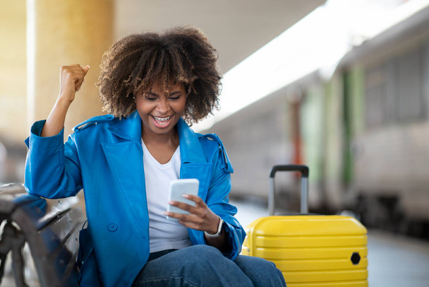 Gioiosa donna nera che festeggia il successo con lo smartphone mentre si siede sul banco alla stazione ferroviaria, Felice afro-americana femminile che gioca giochi online, Divertirsi in attesa di treno, Spazio libero - Foto, immagini