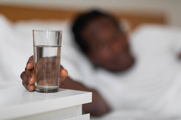 Διψασμένος μαύρος άντρας που παίρνει ένα ποτήρι νερό από το κομοδίνο στην κρεβατοκάμαρα, νεαρός Αφρο-Αμερικανός άντρας που απλώνει το χέρι του σε υγιεινό ποτό αφού ξυπνήσει το πρωί, επιλεκτική εστίαση με ελεύθερο χώρο - Φωτογραφία, εικόνα