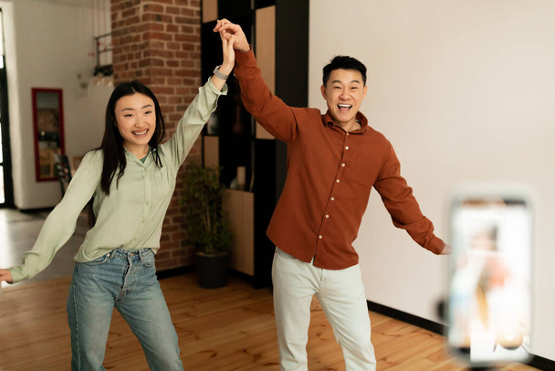 Пара забавных азиатских влиятельных людей снимают танцевальное видео для социальной сети на мобильный телефон, веселятся дома. Мужчины и женщины-блогеры транслируют контент для своего видеоблога, транслируя онлайн - Фото, изображение