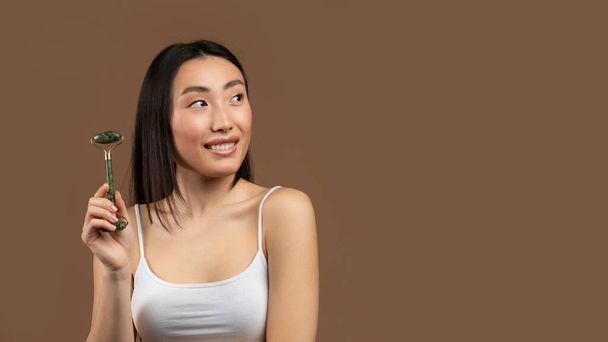 Yeşiltaşı yüz silindiri kullanan Koreli güzel bir bayan kahverengi stüdyo arka planında poz verirken boş alana bakıyor, panorama. Cildiyle ilgilenen Asyalı genç bir kadın. - Fotoğraf, Görsel