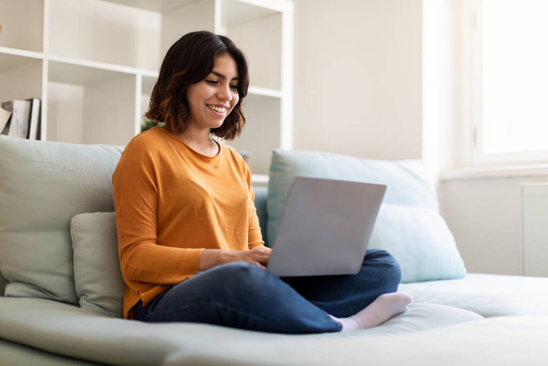 Online Communication. Portrait Of Beautiful Young Arab Woman Using Laptop At Home, Smiling Middle Eastern Female Sitting On Couch In Living Room Interior And Typing On Computer, Copy Space - Foto, Imagen