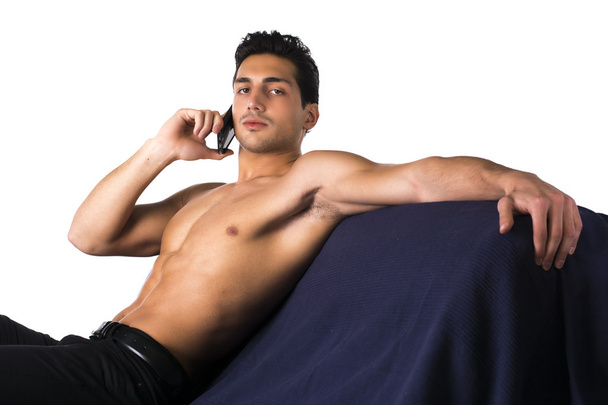 Athletic shirtless young man talking on cell phone sitting on couch - Foto, afbeelding