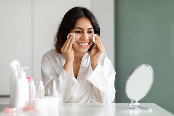 Pretty smiling young middle eastern lady in white silk bathrobe sitting at vanity table full of cosmetics at home, looking at mirror, using cotton pads, removing makeup before sleep, copy space - Foto, Imagen