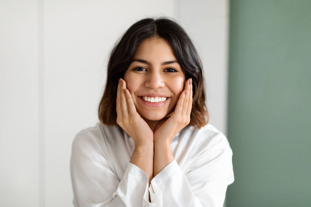 Closeup portrait of cheerful pretty young middle eastern woman in white silk robe touching her face and smiling, enjoying smooth glow moisturised skin, copy space. Face care routine concept - Foto, Imagen