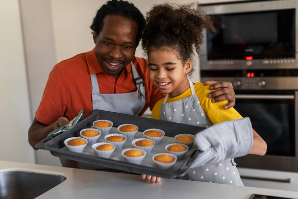 Милая черная девочка и ее отец держат поднос с кексами на кухне, взволнованный африканский американский отец и ребенок-подросток, глядя на свежую выпечку, наслаждаясь приготовлением пищи вместе дома - Фото, изображение
