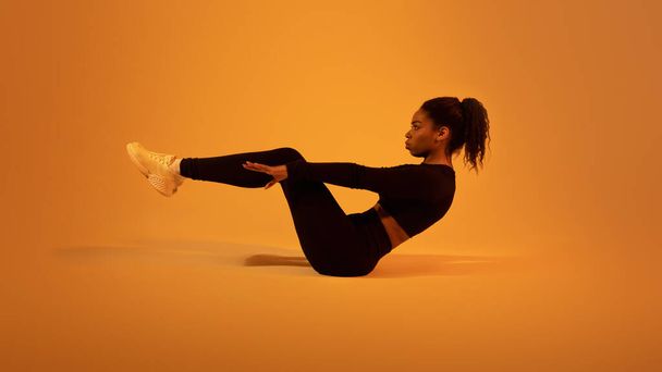 Фитнес-тренировка. Активная чёрная леди делает упражнения на животе, лежащие на полу над оранжевым неоновым студийным фоном, вид сбоку. Решительная женщина с разминающимися мышцами живота - Фото, изображение