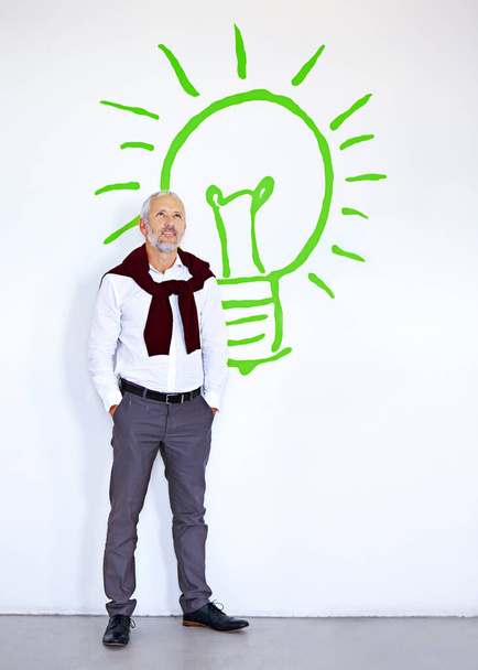 A pensar em novas ideias brilhantes. Um homem de negócios maduro olhando pensativo enquanto estava na frente de uma parede branca com uma lâmpada nele - Foto, Imagem