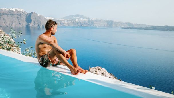 Santorini Greece Oia, young men in swim shorts relaxing in the pool looking out over the caldera of Santorini Island Greece, infinity pool, a young guy on a luxury vacation in Europe Greece - Photo, Image