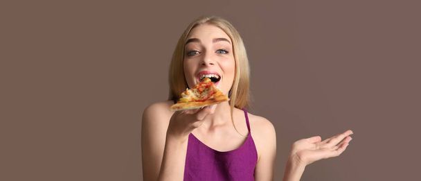 Happy young woman eating slice of tasty pizza on brown background - Photo, Image