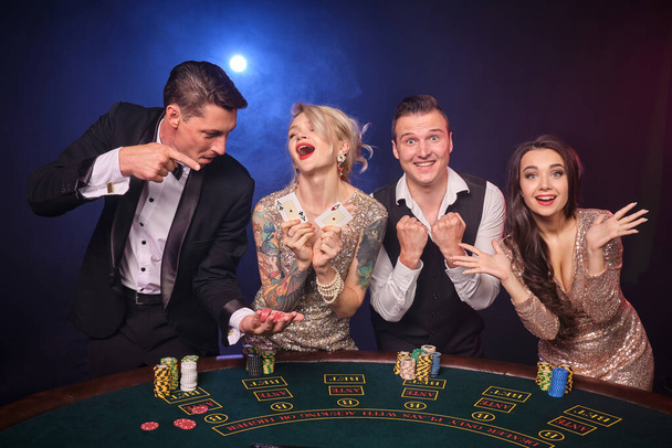 Group of a happy rich buddies are playing poker at casino. Youth are making bets waiting for a big win. They are looking happy standing at the table against a red and blue backlights on black - Foto, imagen