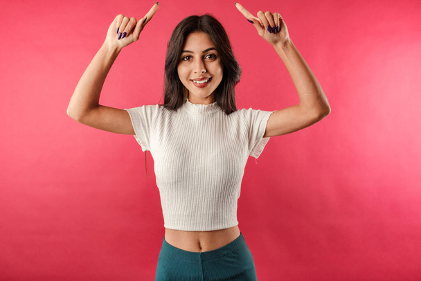 Happy young woman wearing white ribbed crop isolated over red background pointing fingers up at copy space. New product sharing, raise hands pointing fingers up smiling broadly. - Photo, Image