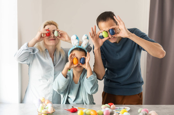 Χαρούμενη οικογένεια που φοράει ακουστικά κουνελιού μαζεύονται στο τραπέζι στη μοντέρνα ελαφριά κουζίνα και βάφουν πασχαλινά αυγά μαζί. - Φωτογραφία, εικόνα