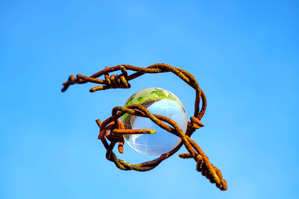 The concept of symbolism. On a blue sky is a glass globe, which is surrounded by rusty barbed wire - Foto, Imagem