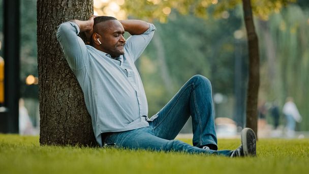 Tranquillo rilassato riposo di mezza età musica di ascolto maschile con auricolari seduti su erba verde nel parco sotto l'albero godere di canzone rilassante uomo etnico afroamericano adulto in cuffie wireless ascoltare audio - Foto, immagini