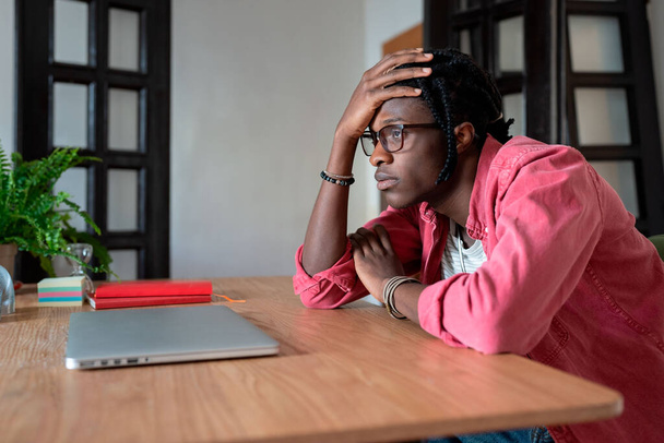 Sad worried young African American man remote worker touching forehead thinking pondering about personal problems at workplace, sitting at desk with closed laptop. Freelancer mental health concept - Foto, Bild