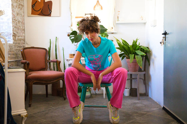 Young and beautiful latina woman dressed in bright colours is sitting on the chair of her fashion design and painting studio posing for the camera doing different postures and expressions. woman power - Photo, Image