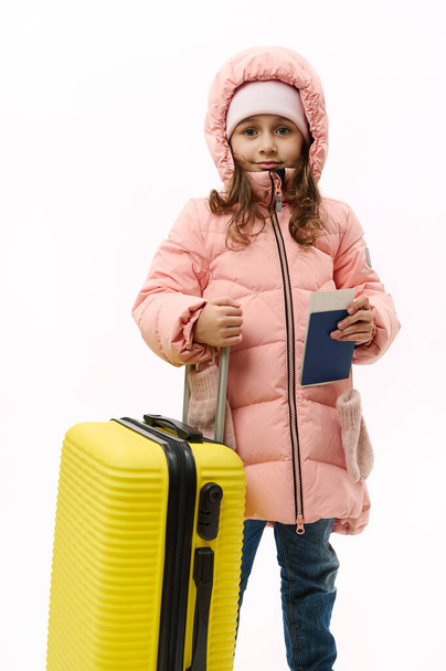 Full-length portrait: Beautiful little girl in pink down coat, going for vacations, looking at camera, posing with suitcase and boarding pass on white background with copy space. Travel Trip Journey - Photo, image