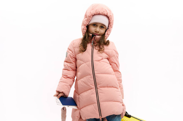 Adorable European child girl in pink down coat, looks at camera, poses with suitcase and boarding pass over white background. Kids going for holidays. air flight. Travel Trip Journey. Winter holidays - Foto, imagen