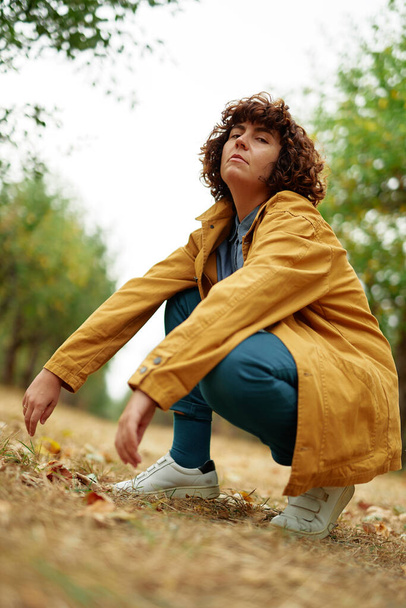 European autumn fall park photo shoot. Autumn portrait of young attractive fashionable sitting curly girl wearing orange jacket looking straight in camera. High quality vertical photo - Foto, imagen