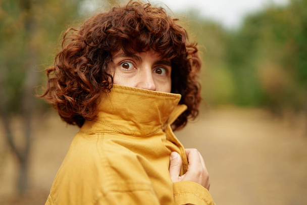 Covering face and making big eyes. Autumn portrait of young attractive fashionable happy curly girl at orange jacket looking straight in camera. European autumn fall park photo shoot - Foto, imagen