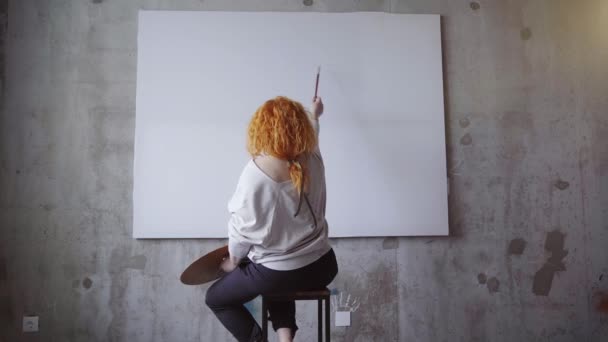 コンクリートの灰色の壁に白い空白のキャンバスの前に座っている陽気な赤毛のアーティストの女性。パレットで傑作を考え、絵画のビジョンを作る女性画家。4kビデオ映像 - 映像、動画