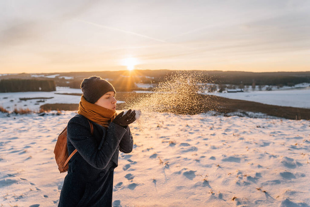 Een vrouw in een jas gewikkeld in een sjaal blaast sneeuw van haar handen. - Foto, afbeelding