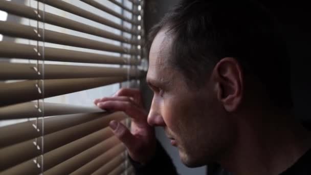 Egy szakállas férfi közeledik az ablakhoz, hátrahúzza a redőnyt, és egy repedésen keresztül néz ki az ablakon. Egy középkorú fehér férfi néz ki az ablakon. - Felvétel, videó