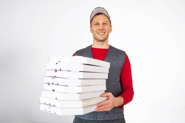 Photo of happy delivery man in red t-shirt and cap giving food order and holding many pizza boxes isolated on white background - Photo, Image