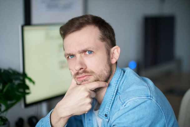 matoucí gesto nebo inspirace koncept: roztomilý vousatý portrét bělocha samec na volné noze softwarový inženýr sedí na pracovišti v kanceláři a dívá se do kamery s počítačem na pozadí - Fotografie, Obrázek