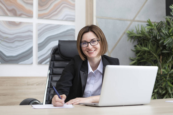 Portrait of successful and attractive business woman working on laptop in her office. Positive female employee in black suit smiling and sitting at the desk in her modern workplace - Foto, Bild