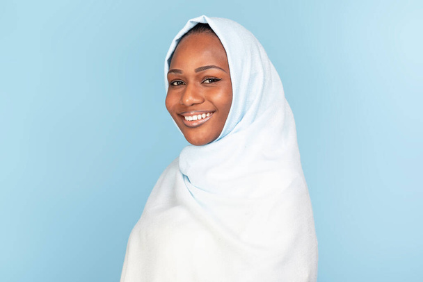 青いスタジオの背景にポーズをとるヒジャーブの美しい黒いイスラム教徒の女性の肖像画、カメラで笑顔、コピースペース。女性の美しさの概念,女性の多様性 - 写真・画像