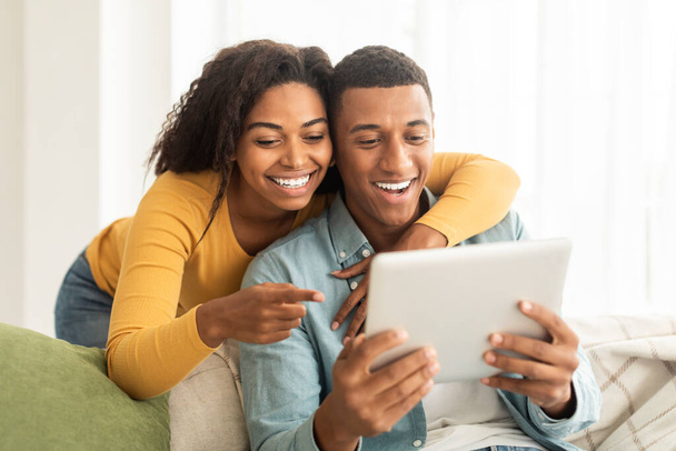 Cheerful millennial african american lady hug guy, looking at tablet, have video call, use app in living room interior, close up. New normal and lifestyle, technology for communication, ad and offer - Photo, image