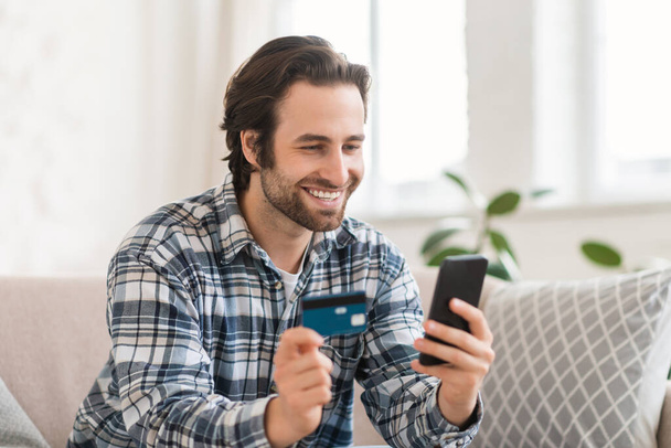 Cheerful young caucasian guy shopaholic sits on sofa with smartphone and credit card, shopping online, checks financial account in room interior. App for banking, sale and ordering goods remotely - Photo, image