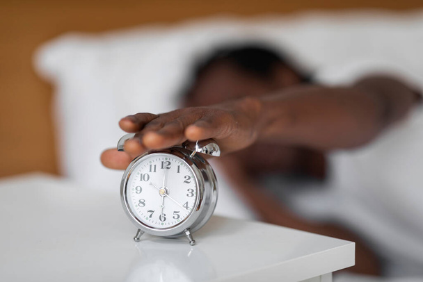 Lähikuva tunnistamaton musta mies saavuttaa herätyskello yöpöydällä käsin, nuori Afrikkalainen amerikkalainen mies makaa mukavassa sängyssä kotona, herääminen nukkumisen jälkeen makuuhuoneessa, valikoiva keskittyä - Valokuva, kuva