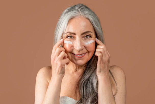 Šťastná starší běloška s přirozenou krásou nanáší na obličej krém, bojuje s vráskami, užívá si anti-aging procedury, izolovaná na hnědém pozadí, zblízka, studio. Kosmetická péče, lázeňská léčba - Fotografie, Obrázek